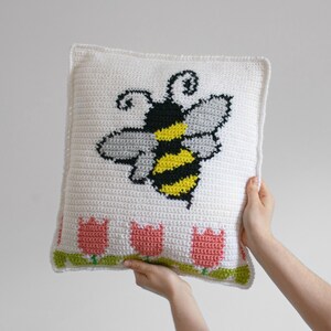 Puede incluir: Un coj&iacute;n blanco tejido a ganchillo con un gran dise&ntilde;o de abeja en negro, amarillo y gris. El coj&iacute;n tambi&eacute;n presenta una fila de tulipanes rosas con tallos y hojas verdes en la parte inferior. El coj&iacute;n est&aacute; sostenido por dos manos.