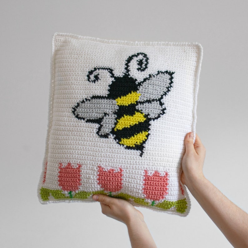 Puede incluir: Un coj&iacute;n blanco tejido a ganchillo con un gran dise&ntilde;o de abeja en negro, amarillo y gris. El coj&iacute;n tambi&eacute;n presenta una fila de tulipanes rosas con tallos y hojas verdes en la parte inferior. El coj&iacute;n est&aacute; sostenido por dos manos.