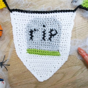 May include: White crocheted Halloween banner with a gray tombstone that says "RIP" in black yarn. The banner has a green border around the tombstone.