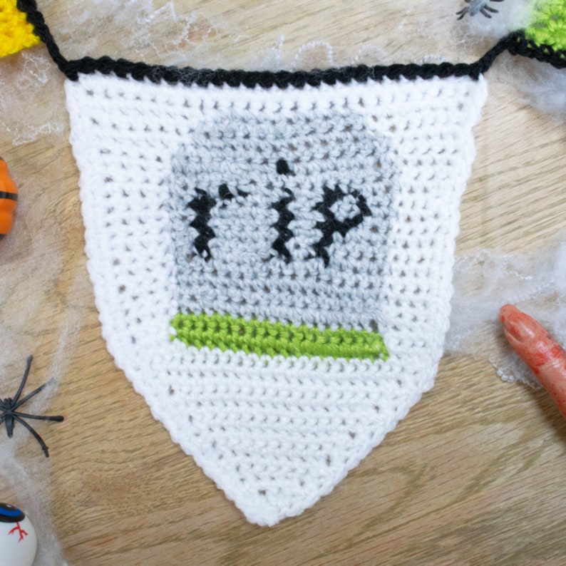 May include: White crocheted Halloween banner with a gray tombstone that says "RIP" in black yarn. The banner has a green border around the tombstone.