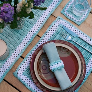 May include: A set of crocheted placemats and a runner in a light blue and white colour scheme. The placemats have a white border and the runner has a white border with a repeating pattern. The placemats are set on a wooden table with a plate, fork, knife, and napkin.