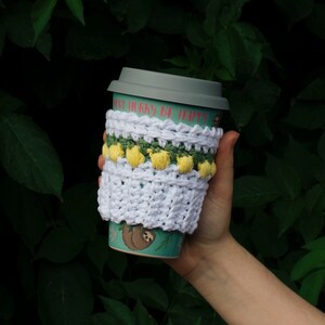 May include: White crocheted coffee cup cozy with yellow and green accents. The cozy has a scalloped edge and a small sloth design on the cup. The text "Don't hurry be happy" is printed on the cup.