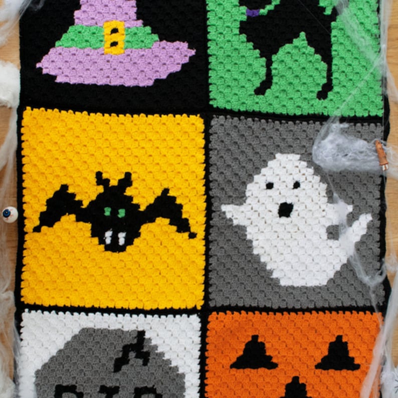 May include: A colorful crochet Halloween blanket with squares featuring a witch's hat, a bat, a ghost, a black cat, and a jack-o'-lantern.