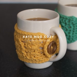 May include: A white ceramic mug with a yellow crocheted cosy and a wooden button closure. The mug contains a warm beverage. Text on the cosy reads "KATE MUG COZY" and "sunflower cottage crochet". Another mug with a green cosy is in the background.