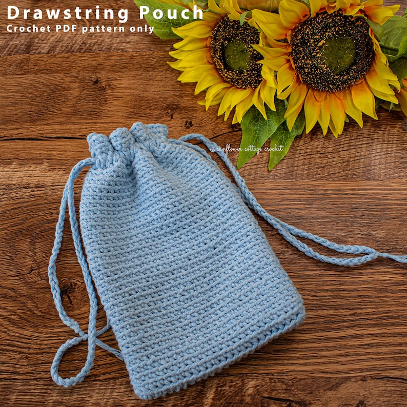 May include: A light blue crocheted drawstring pouch with a white drawstring. The pouch is lying on a wooden surface with sunflowers in the background.
