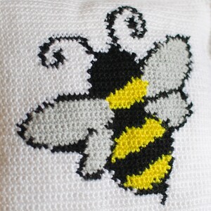 Puede incluir: Un coj&iacute;n blanco con un dise&ntilde;o de punto de cruz de una abeja. La abeja tiene rayas amarillas y negras en su cuerpo, alas grises y antenas negras. El dise&ntilde;o est&aacute; centrado en el coj&iacute;n.