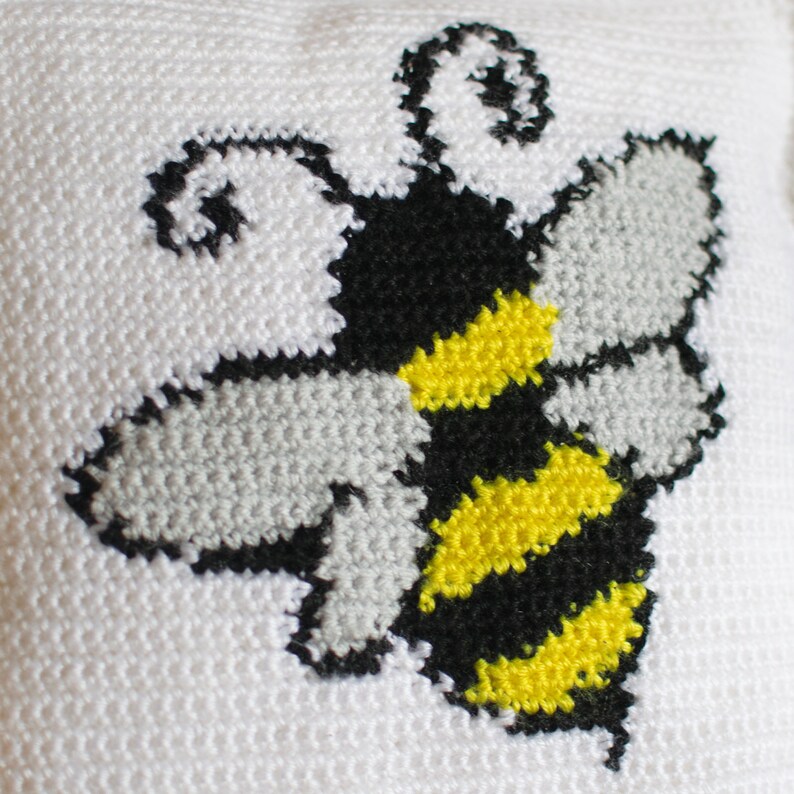 Puede incluir: Un coj&iacute;n blanco con un dise&ntilde;o de punto de cruz de una abeja. La abeja tiene rayas amarillas y negras en su cuerpo, alas grises y antenas negras. El dise&ntilde;o est&aacute; centrado en el coj&iacute;n.