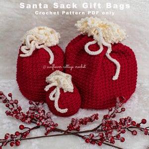 Könnte beinhalten: Drei rote gehäkelte Santa-Sack-Geschenktüten in verschiedenen Größen, mit cremefarbenen Oberteilen und Kordelzügen. Die Taschen sind auf einer weißen Oberfläche mit roten Beerenzweigen angeordnet. Der Text oben lautet "Santa Sack Gift Bags Crochet Pattern PDF only".
