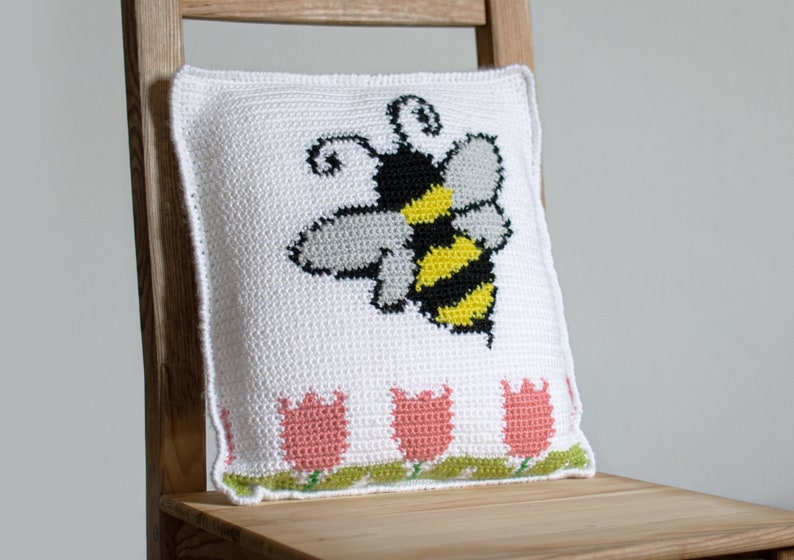 Puede incluir: Coj&iacute;n cuadrado blanco con un dise&ntilde;o de abeja de ganchillo en negro, amarillo y gris. El borde inferior presenta dise&ntilde;os de tulipanes rosas con hojas verdes. El coj&iacute;n est&aacute; sobre una silla de madera.