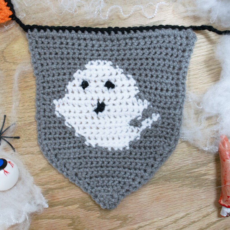 May include: A gray crocheted Halloween banner with a white ghost in the center. The ghost has two black eyes and a black mouth.