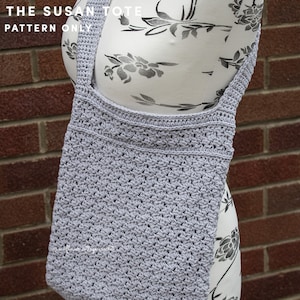May include: A gray crocheted tote bag with a wide strap. The bag is hanging on a white dress form with a floral pattern. The text "THE SUSAN TOTE PATTERN ONLY" is at the top of the image.