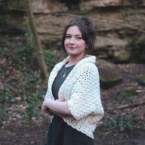 Peut inclure: Une femme portant un cardigan en crochet blanc avec un chemisier vert en dessous. Elle se tient dans une zone boisée avec un fond rocheux.