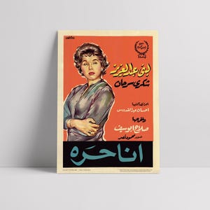 May include: Vintage movie poster featuring a woman in a gray blouse against an orange background. The poster has Arabic text and a black banner at the bottom with more text. The poster is displayed on a white surface.