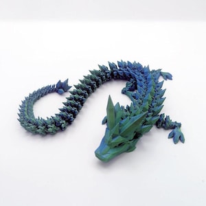 Op de afbeelding: Een 3D-geprinte drakenfiguur in tinten blauw en groen. Het lichaam van de draak is gesegmenteerd, met een kristalachtige textuur. De kop en staart zijn gedetailleerd met scherpe, puntige kenmerken, wat het een fantasie-esthetiek geeft.
