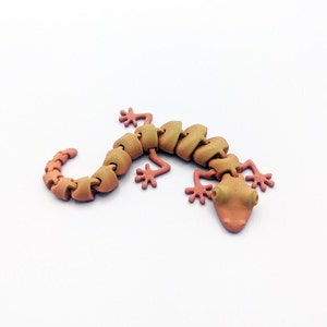 May include: A segmented, articulated, 3D printed gecko lizard toy with a light brown body and pink limbs. The gecko is posed in a walking position with its tail curled.
