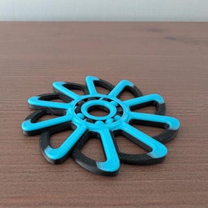 以下が含まれることがあります： 花のようなデザインの3Dプリントされたフィジェットスピナー。スピナーは、8つの湾曲した花びらと黒い外側のリングを備えた明るい青色のフレームが特徴です。中央には回転用のボールベアリングが含まれています。