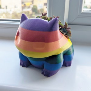 May include: A rainbow-colored 3D printed planter shaped like a Pokemon character. The planter has a purple, red, orange, yellow, green, blue, and purple striped design. The planter is filled with small green plants.