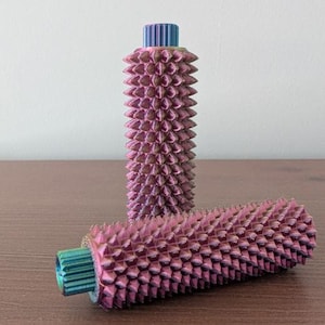 May include: Two decorative cylinders with a textured, spiky surface and an iridescent finish. One stands vertically, the other lies horizontally. The cylinders display a gradient of pink, purple, and blue hues, set against a neutral backdrop.