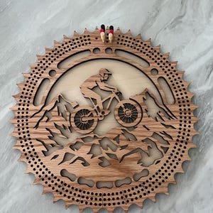 Peut inclure: Un plateau de cribbage circulaire en bois en forme de pignon de vélo, présentant une scène de VTT découpée au laser avec un cycliste sur un sentier. Le plateau comprend des rangées de trous pour le score et quatre petits pions en bois rouge et noir.