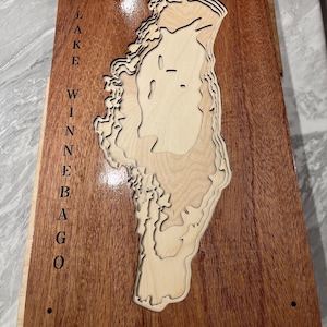 May include: Wooden Lake Winnebago map with layered topographic design. The map is made of light-colored wood and is mounted on a dark brown wooden base. The name "LAKE WINNEBAGO" is vertically engraved on the left side. The map is a decorative piece.