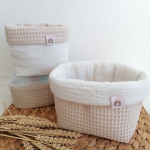 Könnte beinhalten: Drei beige-weiße Stoff-Aufbewahrungskörbe mit Waffelstruktur. Jeder Korb hat eine weiße Innenseite und ein kleines Etikett mit Regenbogen-Design. Die Körbe sind auf einer gewebten Matte mit Weizenähren angeordnet.