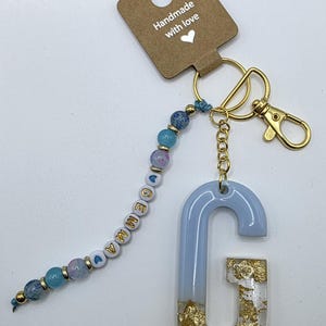 Puede incluir: Un llavero hecho a mano con una letra "G" de resina azul claro y detalles de pan de oro. El llavero incluye un dije de cuentas con el nombre "Gemma" en letras doradas y un cierre dorado. La etiqueta dice "Hecho a mano con amor".