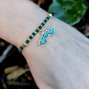 Op de afbeelding: Een kralenarmband met een gouden vleermuisbedel. De armband heeft zwarte, groene en gouden kralen. De vleermuisbedel is groen met een gouden omtrek. De armband wordt om een pols gedragen.