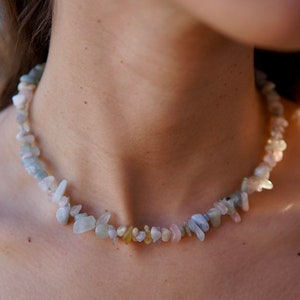 Peut inclure: Gros plan d'une personne portant un débardeur blanc avec un collier délicat composé de petites pierres irrégulières bleu clair, rose et jaunes.