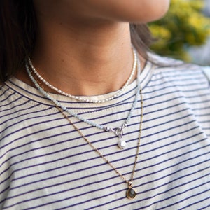 以下が含まれることがあります： 真珠と涙型のペンダントが付いた重ね付けネックレス。ネックレスはシルバーとゴールドのチェーンで作られており、ライトブルーとホワイトのビーズが特徴です。