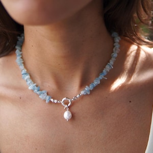 以下が含まれることがあります： 薄い水色のアクアマリンの石と、単一の真珠のペンダントで作られた繊細なネックレス。ネックレスは銀の留め金でできています。