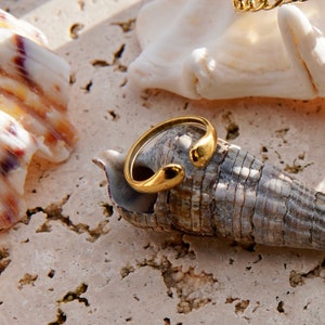 Könnte beinhalten: Ein goldener Ring mit einem einzigartigen, geschwungenen Design. Der Ring liegt auf einer Muschel.