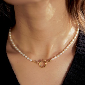 Könnte beinhalten: Eine zarte Perlenkette mit einem goldenen Verschluss. Die Kette besteht aus kleinen, weißen Perlen und hat ein schlichtes, elegantes Design.