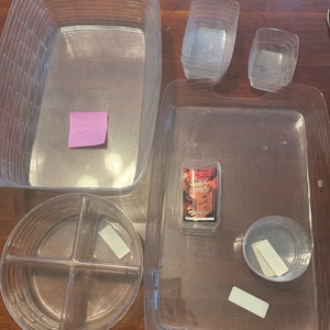 Könnte beinhalten: Ein Satz durchsichtiger Plastik-Aufbewahrungsbehälter mit Deckeln. Die Behälter sind gestapelt und auf einer Holzoberfläche angeordnet. Die Behälter sind in verschiedenen Größen und Formen, darunter ein rechteckiger Behälter, ein runder Behälter mit vier Fächern und ein quadratischer Behälter. Die Behälter sind aus klarem Kunststoff und haben eine glatte Oberfläche.
