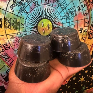 May include: Four black circular soap bars stacked on top of each other. The soap bars are on a hand, which is holding them over a colorful tapestry with a zodiac wheel design.
