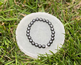 May include: A black hematite beaded bracelet with a silver clasp. The bracelet is on a white selenite charging plate.