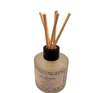 May include: A frosted glass reed diffuser with a black cap and wooden reeds. The label reads "Anna Elizabeth Candles" and "Fairy Dust". The diffuser is designed to release fragrance into the air.