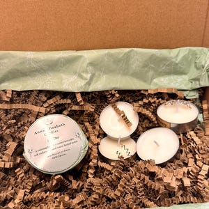 May include: A gift box with candles and decorative paper. The box is brown with the text "Thoughtful scents for everyday moments." Inside are several small, round candles in metal holders, and a round candle with a label. The box is filled with brown paper.