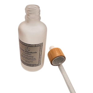May include: A frosted glass bottle with a dropper. The bottle has a label that reads "Anna Elizabeth Candles Frosty Mornings". The dropper has a wooden top and a clear glass tube. The bottle is empty.