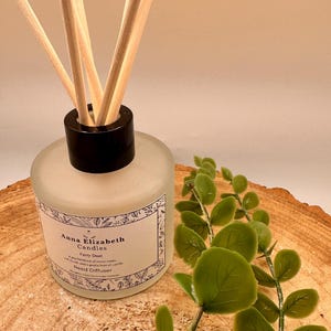 May include: A frosted glass reed diffuser with a black cap and wooden reeds. The label reads "Anna Elizabeth Candles Fairy Dust". Green leafy sprigs are placed next to the diffuser on a wooden surface.