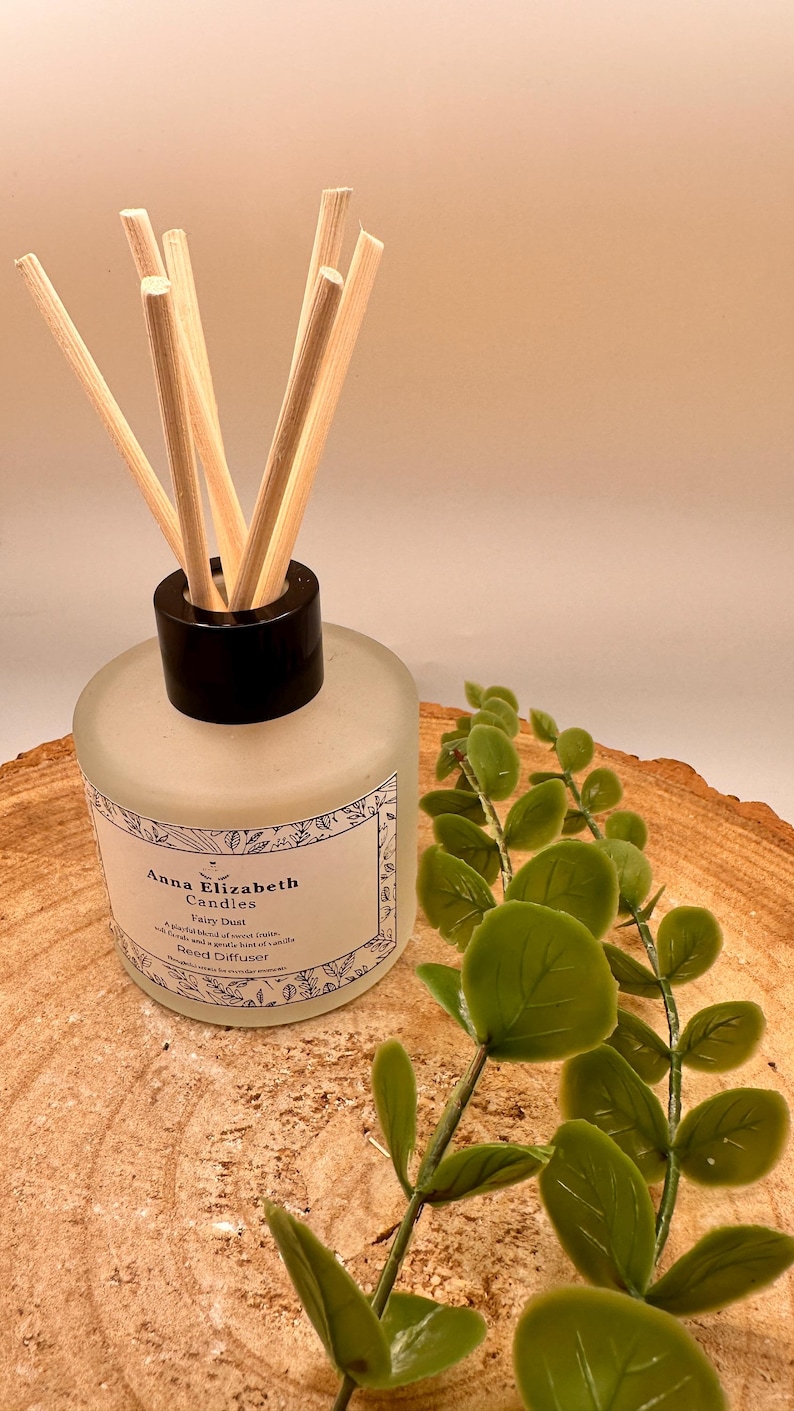 May include: A frosted glass reed diffuser with a black cap and wooden reeds. The label reads "Anna Elizabeth Candles Fairy Dust". Green leafy sprigs are placed next to the diffuser on a wooden surface.