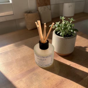 May include: A clear glass diffuser bottle with a black cap and wooden reeds. The bottle has a label that reads "Anna Elizabeth Candles". A small potted plant sits next to the diffuser on a wooden surface. The overall theme is home fragrance and decor.