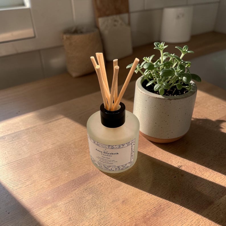 May include: A clear glass diffuser bottle with a black cap and wooden reeds. The bottle has a label that reads "Anna Elizabeth Candles". A small potted plant sits next to the diffuser on a wooden surface. The overall theme is home fragrance and decor.
