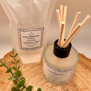 May include: A clear pouch and a frosted glass bottle with reed diffuser sticks. Both feature labels with the text "Anna Elizabeth Candles Coastal Walks". The pouch has a white cap. Green leafy sprigs are in the foreground.