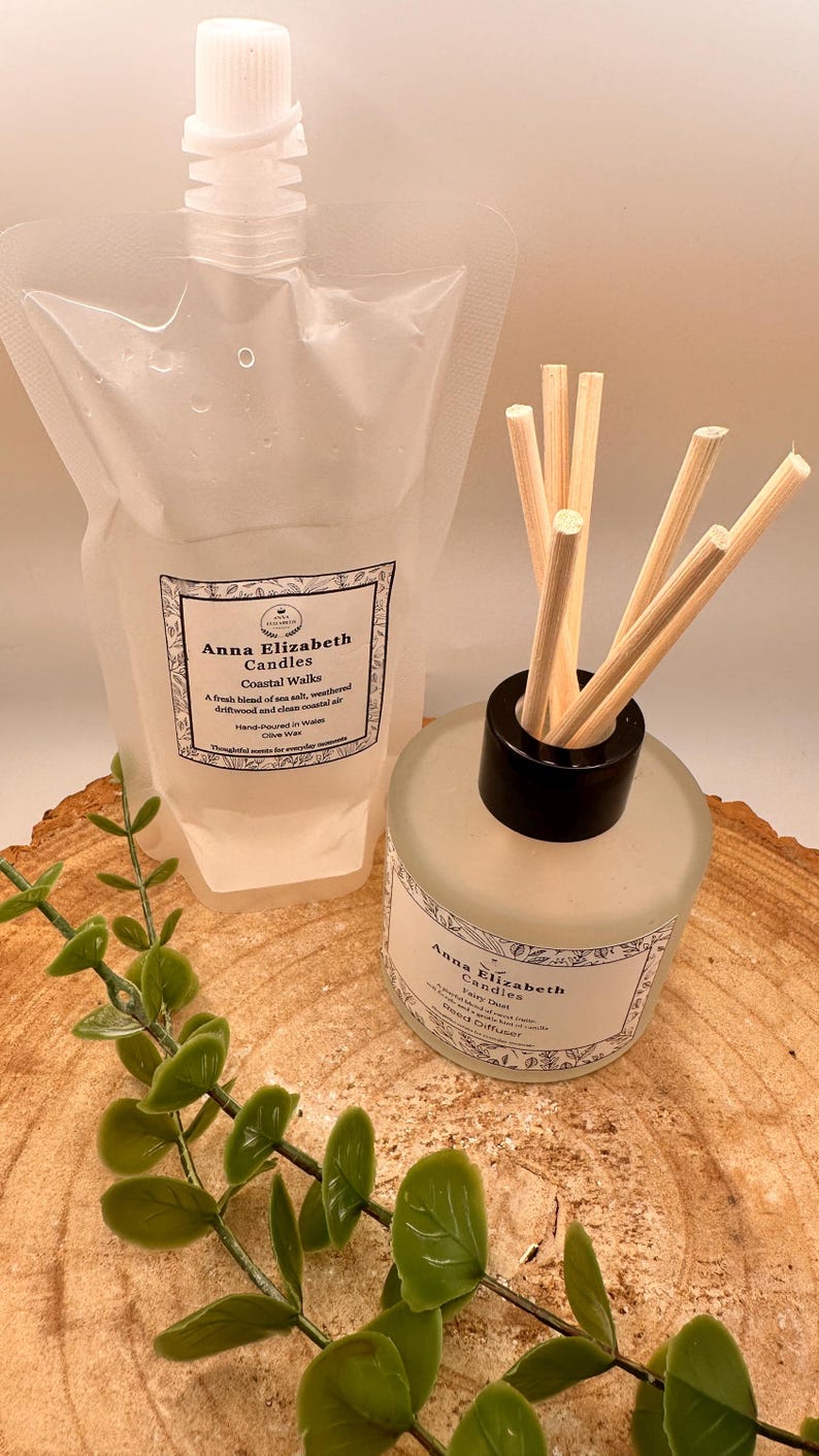 May include: A clear pouch and a frosted glass bottle with reed diffuser sticks. Both feature labels with the text "Anna Elizabeth Candles Coastal Walks". The pouch has a white cap. Green leafy sprigs are in the foreground.