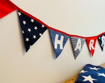 May include: A red, white, and blue fabric banner with the word "HAPPY" spelled out in white letters. The banner is strung up on a white wall.