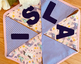 Könnte beinhalten: Ein lila-rosa Stoffbanner mit weißen Punkten und einem Einhorn-Druck. Das Banner hat die Buchstaben "ISLA" in schwarzem Filz.