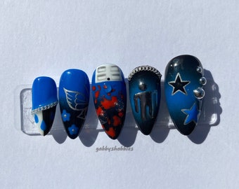 May include: Set of six blue and black press-on nails with various designs, including a microphone, a butterfly, a red and black splatter, a stick figure, and a star. The nails are displayed on a white card with the text "gabby's hobbies".