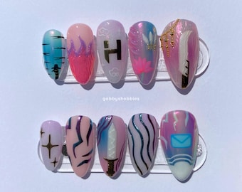 Pode incluir: Um conjunto de unhas postiças com vários designs. As unhas apresentam uma variedade de cores, incluindo azul, rosa e roxo, com padrões como chamas, listras e motivos florais. Algumas unhas têm a letra "H" ou um design de envelope. O texto "gabby's hobbies" é visível.