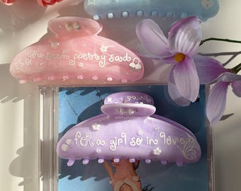 May include: Three hair claw clips in light blue, pink, and purple. The blue and pink clips feature the text "you seem pretty sad" with small star, bow, and padlock icons. The purple clip features the text "for a girl so in love" with flower and butterfly icons.