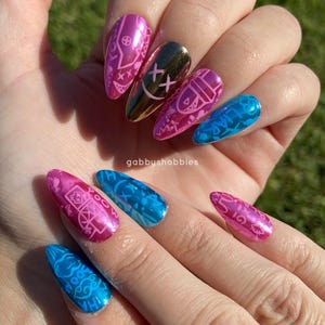 May include: A hand displaying a set of long, almond-shaped artificial nails. The nails feature a variety of designs, including pink and blue metallic finishes with intricate patterns and a gold nail with a black smiley face design.